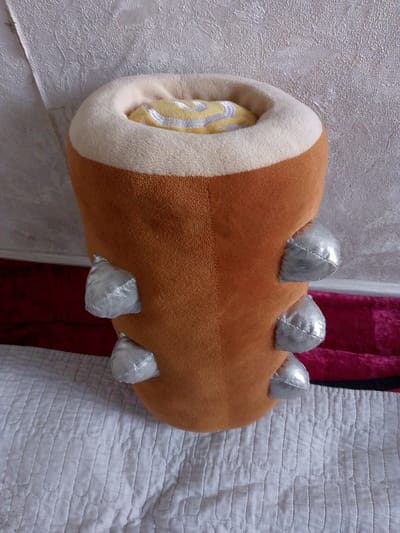 A plush toy resembling a brown barrel with a cream-colored rim and silver, cone-shaped protrusions is positioned upright on a white quilted surface. Inside the rim, a yellow and white striped fabric is visible. The toy is situated against a textured, light-colored wall. The background also includes a dark red surface and a bright pink fabric. The image is taken from a slightly elevated angle, looking down at the toy. The setting appears to be indoors, possibly a room. There are no people visible in the image.