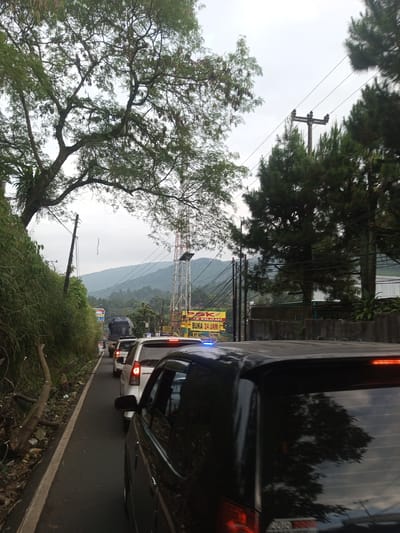 A street in Taunggyi, Myanmar, is congested with cars in a traffic jam. The vehicles are primarily cars, including a black van in the foreground with its brake lights illuminated. On the left is a roadside covered with foliage and a fallen tree limb. A tall telecommunications tower and mountains are visible in the background under a cloudy sky. An overhead power line follows the street. A sign with yellow lettering that reads "BUKA 24 JAM" is also visible in the background.