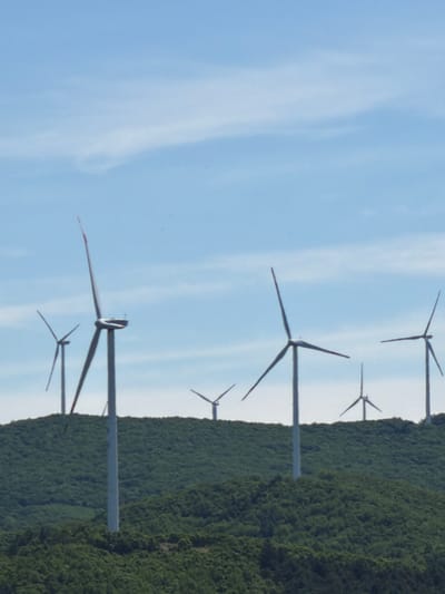 The image, taken in Jiejinkou, China, depicts a landscape dominated by a wind farm. The scene is composed of a clear blue sky on the left and a lush, green, forested hillside on the right, separated by a distinct horizon line that runs vertically through the image due to its orientation.

Multiple wind turbines are visible, distributed across the frame. There are six distinct turbines, varying in proximity and size, suggesting they extend into the distance. The closest turbine is prominent, with its tall white tower and three dark blades clearly visible against the blue sky. Its blades appear stationary, some showing subtle red markings near their tips. Further back, other turbines are partially obscured by the foreground turbine or appear smaller, receding into the background.

The forested hillside is densely covered with trees, indicating a natural or reforested area, providing a natural contrast to the industrial structures of the wind farm. The sky is bright blue with faint, wispy white clouds scattered across it, suggesting a sunny daytime. There are no people or visible text in the image. The overall impression is one of renewable energy integration into a natural, hilly environment.