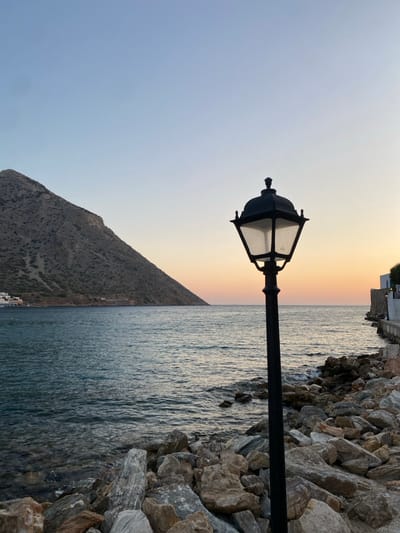 The image depicts a coastal scene at dusk or dawn in Apollonia, Greece, viewed from a rocky shoreline. A prominent black, unlit, ornate street lamp with four clear glass panels stands vertically in the right-center foreground, positioned among large, irregularly shaped gray and brown rocks that form the immediate coastline. The sea, a dark blue-green, stretches across the midground with visible surface ripples and small waves breaking against the foreground rocks. To the left, a large, steep, barren mountain with sparse vegetation rises directly from the water, dominating the left side of the frame. At the far left base of this mountain, a small cluster of white-washed buildings with blue elements is visible near the water's edge. Along the distant right shoreline, a stone wall and additional white buildings are discernible. The sky transitions from light blue at the zenith to soft orange and pink hues at the horizon. No individuals or active subjects are present.