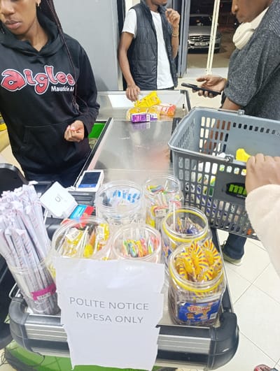 A cashier in a black hoodie stands at a checkout counter. A man in a black vest is next in line, followed by another man holding a phone and a shopping basket. The counter displays items being purchased, including bread and other groceries. Candy in plastic containers and paper straws fill the front display. A sign reads "POLITE NOTICE MPESA ONLY," indicating a payment preference.