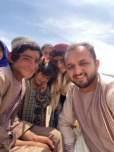 A high-angle, outdoor snapshot shows seven people, apparently in a sunny locale, likely in the city of Ashoqah, Afghanistan. The foreground is occupied by three men. The man on the left is wearing a tan jacket over a patterned shirt and a crocheted cap. Next to him and slightly behind is a young girl in a green patterned dress and necklace. To the right is a man wearing a shirt with a brown and white embroidered pattern. All three are smiling into the camera. Behind them are four more people, partially obscured. A girl in a blue headscarf is visible on the far left. Two other girls are partially visible in the center of the frame, and a boy in a red cap is also smiling into the camera. The background reveals a light blue sky with some thin clouds. The lighting is bright, suggesting a daytime setting.