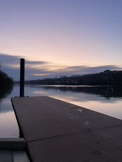 A lakeside dusk scene displays a wooden dock extending into still water. A dark post anchors the dock. Across the water, faint city lights glimmer against a backdrop of shadowy hills. The sky above is a blend of pale purples and yellows, reflecting in the water's surface. The dock's planked texture and the smooth water contrast the natural, silhouetted landscape.