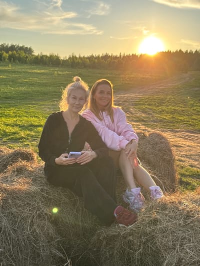 The image displays two women seated outdoors during sunset in a rural or pastoral setting, identified as Стеклянный, Russia. The photograph is oriented as if the sun is on the left, casting a strong golden backlight across the scene.

The setting sun is visible on the left side of the frame, low on the horizon behind a line of trees, creating a bright glow and warm light. The sky has some wispy clouds. The ground where the women are sitting is covered in dry grass or hay, with a noticeable hay bale partly visible behind one of them. In the background, a field of green grass or crops stretches towards the tree line.

One woman, with light brown hair, is wearing a light pink long-sleeved jacket or top. She smiles warmly at the camera, her legs extended, wearing light-colored athletic shoes (one purple, one reddish-purple). She appears to be leaning against or sitting on a mound of hay.

The second woman, with blonde hair tied back, is wearing a dark, possibly black, long-sleeved zipped top and dark pants. She is also smiling at the camera, holding a light-colored smartphone in her hands. A small, glowing yellow-green object is discernible on the dry grass near her leg.

The women are engaged in the activity of posing for a photograph. Their smiles convey a relaxed and joyful emotion. The time of day is clearly sunset, characterized by the low sun and golden hour lighting. No visible text is present in the image.