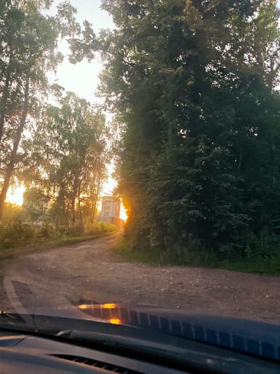 The image depicts a dirt road winding through a wooded area in Snt Vodnik, Russia, during what appears to be sunset or sunrise. Tall trees with dense foliage flank the road, with the sun's rays filtering through the leaves, creating a warm, golden glow. On the left side of the road, between the trees, a building with multiple windows and a light-colored facade is visible. Further down the road, another building, possibly an apartment complex, can be seen with a similar architectural style. The sky is bright, suggesting a clear weather condition. The foreground shows the dashboard and windshield of a vehicle, reflecting the ambient light and the road ahead. The overall mood is serene and peaceful, evoking a sense of a quiet rural journey. There is no visible text in the image.