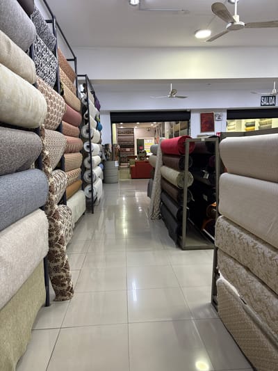 The image depicts the interior of a fabric store in La Victoria, Peru. The store is filled with large rolls of fabric displayed on metal shelving units that line the aisles. The fabrics vary in color, pattern, and texture, ranging from neutral tones like beige and grey to more vibrant hues like red and purple. Some rolls are plain, while others feature intricate floral or geometric designs. In the background, a sign reads "SALIDA," indicating an exit. Two ceiling fans are visible, suggesting the environment is warm. The floor is tiled with a reflective surface, creating a polished appearance. The overall scene is one of an organized retail space dedicated to the sale of textiles.