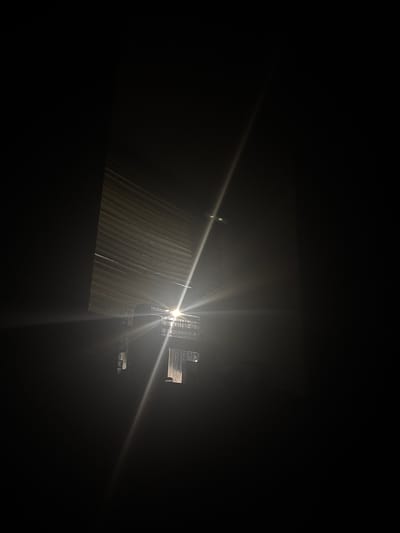 The image depicts a nighttime scene in Welshpool, United Kingdom. Light emanates from what appears to be a building with corrugated metal siding. The light source is bright, causing a lens flare effect. The environment is dark, obscuring details outside of the immediate area illuminated by the light. There are no visible people or interactions in the image.