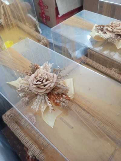 Three clear, rectangular boxes are arranged. Each box contains dried wheat stalks, and a large beige flower with a small ribbon embellishes the corner. The box in the foreground has a prominent flower and a woven basket underneath. Another box has a similar floral arrangement, and the third one is partially visible, showing its contents and adjacent items. A red and white box is also present in the background.