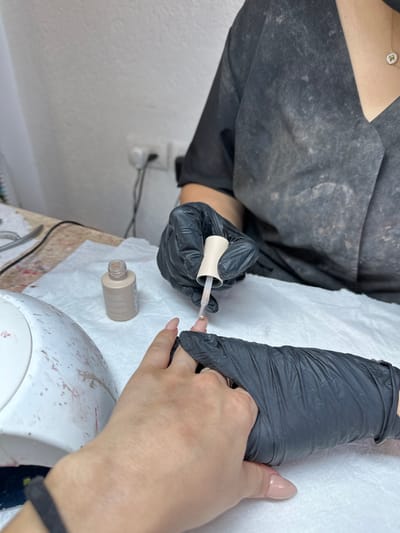 A close-up, high-angle interior image captured in Kafr Kasim, Israel, depicts a manicure in progress. A nail technician, wearing a dark, possibly stained, short-sleeved smock and black protective gloves, is applying a substance to a client's right hand. The technician's gloved right hand holds a light-colored nail polish brush over the client's ring finger, while the gloved left hand supports the client's right hand. The client's right hand features elongated, oval-shaped nails with a light nude or clear coating, and a silver-toned ring is visible on the fourth finger. A black wristband is partially visible on the client's lower left wrist. On the white disposable mat covering the light-colored table, a small, light brown/nude nail polish bottle is positioned near a white UV/LED nail lamp, which displays visible polish stains. The background reveals a plain white wall with a white electrical outlet and plug, while various metallic manicure tools are partially visible on the table's left edge.