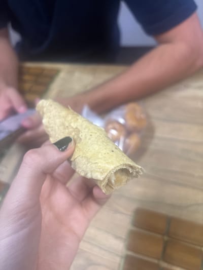 A person holds a partially eaten, rolled taco (flauta) in their right hand. The fingernails on the right hand are painted dark green. A second person, wearing a dark blue shirt, is partially visible to the left of the frame. In the background, there are cylindrical objects, possibly tamales, stacked in rows, and a plastic bag containing round objects, possibly donuts or rolls. The setting appears to be indoors, on a wooden surface. There is no information to deduce a specific location in Buenavista, Mexico.