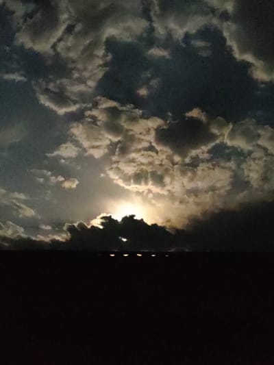 This is a nighttime photograph taken from an elevated vantage point overlooking the city of Qargaly, Kazakhstan. The sky is filled with dramatic, dark clouds illuminated from behind by a bright, unseen light source, possibly the moon or artificial lighting. The clouds are textured with highlights and shadows, suggesting movement and depth. Below the sky, a dark, silhouetted horizontal line likely represents the cityscape or a rooftop. A series of small, evenly spaced, glowing orbs are visible along this line, suggesting streetlights or building lights. The lower portion of the image is almost entirely black, indicating a deep shadow or darkness. The overall atmosphere is dramatic and moody.