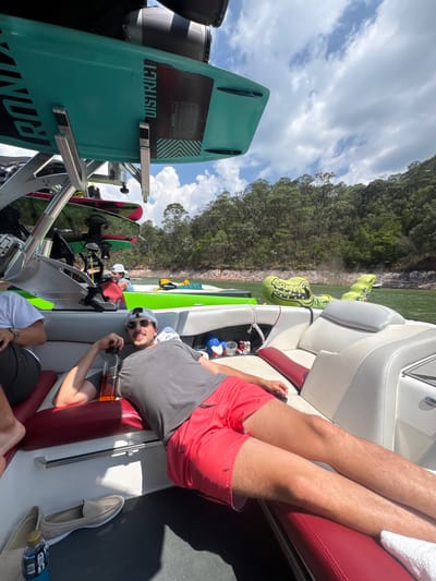 The image depicts a group of people relaxing on a boat on a lake in Valle de Bravo, Mexico. The boat is equipped with wakeboards and an inflatable alligator float. A man in red shorts and a gray shirt is lying on the boat's seating, holding a bottle. The scene includes elements such as trees, water, and skies, indicative of a sunny day on the lake.