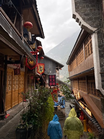 The image depicts a narrow, stone-paved alleyway descending through the historic city of Gongtan, China, under an overcast sky. Two individuals, seen from behind, walk down the path wearing hooded rain ponchos; one is light blue and the other yellow-green.

On the left, multi-story traditional Chinese wooden buildings feature dark tiled roofs and prominent balconies. Red Chinese lanterns hang from the eaves and balconies, interspersed with red and yellow banners displaying Chinese characters, including "红红火火烧烤" (Hónghónghuǒhuǒ Shāokǎo - "Prosperous/Red Hot BBQ"). Clothes are visible hanging to dry on an upper balcony. The ground floor exhibits finely carved wooden doors and shutters. A red and yellow broom rests against the building near the pathway. Lush green foliage, including potted plants, lines the left side of the path.

On the right, another multi-story building displays traditional architecture with a stone base and wooden upper sections featuring numerous lattice-patterned window frames. An illuminated section with warm light is visible below these windows, possibly a shopfront.

In the background, a large mountain shrouded in mist rises under the cloudy sky, dominating the vista between the buildings. The pathway consists of irregular stone slabs and steps.
