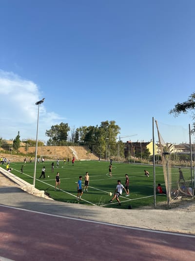 The image depicts an outdoor, artificial turf soccer field in Fuengirola, Spain. Several individuals, predominantly young males, are engaged in a casual soccer game or practice session. Some are kicking or dribbling balls, while others are observing or resting. The field is marked with white lines. In the background, there are trees, buildings, and a construction crane, suggesting an urban or semi-urban environment. A chain-link fence with netting encloses part of the field. A lamppost with multiple light fixtures is positioned on the left side of the frame. The sky is clear and blue with some wispy clouds. A red running track is visible in the foreground.