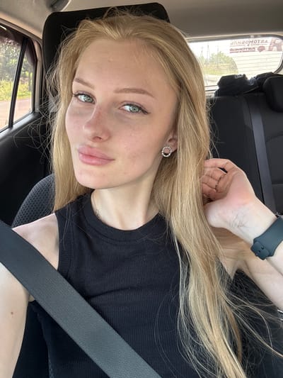 A young blonde woman with green eyes and light skin is in the passenger seat of a car. She is wearing a black tank top and a seatbelt. Her hair is long and blonde, and she is wearing earrings and a watch. She is looking at the camera and has her hand on her hair. The car appears to be parked, with trees and buildings visible through the windows. The license plate visible on the rear window has Cyrillic text that reads "КРАСНЫЙ ВОСХОД".