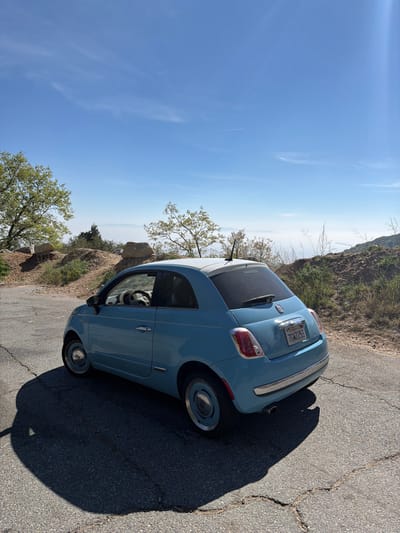 A light blue Fiat 500 compact car is parked on an asphalt road in a mountainous area near Crestline, California, United States. The vehicle, featuring a white roof, chrome trim accents, and light blue hubcaps, displays a California license plate number 7GWX253 on its rear. The interior, including a dark steering wheel, is visible through the open driver's side window. The asphalt road surface exhibits multiple cracks. To the left of the car, a dirt slope with exposed rocks and sparse green trees and shrubs ascends. To the right, the terrain slopes upward with dry grasses and scattered vegetation. The distant background reveals a clear blue sky with a faint aircraft contrail, transitioning to a broad layer of atmospheric haze or fog obscuring the lower landscape, indicating a high elevation vantage point. No individuals or other active subjects are present in the scene. The car casts a distinct shadow to its left, suggesting bright sunlight.