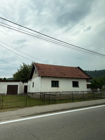 A white house with a red tile roof and two dark windows is situated behind a decorative metal fence. To the left of the house is a smaller white building with a large brown garage door. Power lines are strung across the cloudy sky above. The scene is viewed from a street with a white line marking the edge of the road. The location is identified as Rastovci, Bosnia and Herzegovina.