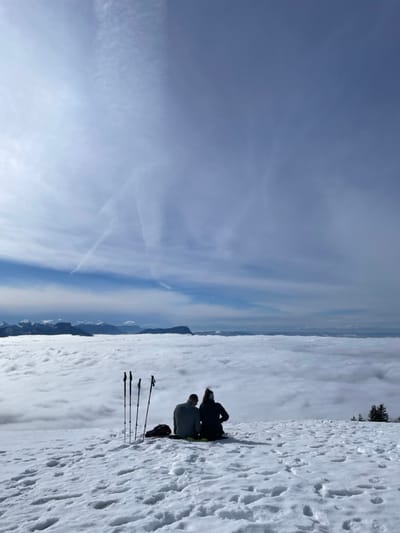 A high-altitude winter mountain landscape is depicted, located in the mountainous region near Les Déserts, France. Two individuals, viewed from behind, are seated directly on a snow-covered ridge, facing a vast cloud inversion that extends to a distant mountain horizon. The person on the left wears a grey long-sleeved top and dark trousers. The person on the right wears a dark jacket, dark trousers, and a light-colored beanie. Four dark ski or hiking poles are upright in the snow to the left of the individuals, adjacent to a dark backpack. The snow-covered ground displays varied textures. The sky above is predominantly clear blue with scattered cirrus clouds and visible contrails. Several conifer trees are present on the far right of the snowline.