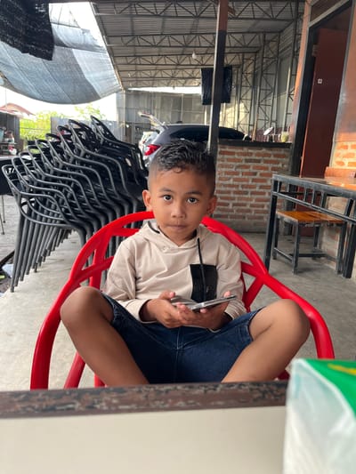 A young boy sits in a red plastic chair, holding a smartphone. He is wearing a light-colored long-sleeved shirt and denim shorts. The boy is looking directly at the camera with a neutral expression. He is in an outdoor setting, with a stack of black chairs behind him. The background includes a corrugated metal roof supported by metal beams, brick walls, and some greenery. A white table is partially visible in the foreground. The scene appears to be a casual, possibly outdoor dining or seating area. The lighting suggests it is daytime. There is no visible text in the image. The location context is Tambonbaroh, Indonesia.