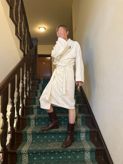 An individual with short, light-colored hair stands on a carpeted staircase within Kliczków Castle in Kliczków, Poland. The subject wears a white terrycloth bathrobe, secured at the waist with a tie. A small green tree logo is visible on the left chest area of the bathrobe. Their right hand is positioned over their face, obscuring facial features, while their left hand holds a set of keys with a light blue key fob. The subject wears tall, dark brown leather boots on both feet. The staircase features dark wooden banisters and balusters. The stair treads are covered with a dark green carpet featuring an ornate pattern of lighter green and white foliage, with a large "K" emblem visible on some steps. In the background, a wooden door with a brass door plate is visible, and an overhead light fixture illuminates the scene from the ceiling.