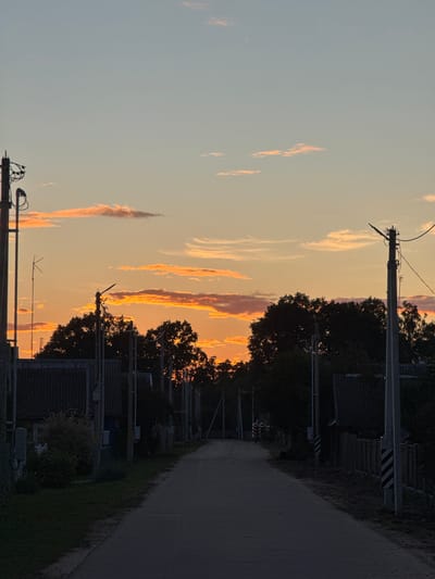 The image depicts a rural road in the Neman Rural Council, Belarus, at sunset. The sky is a gradient of pale blue at the top, transitioning to orange and pink hues near the horizon, with scattered wispy clouds catching the last light. Silhouetted against the vibrant sky are the dark shapes of trees and buildings, including houses with corrugated roofs. Utility poles with electrical wires and what appear to be antennas are visible along the sides of the road. The road itself is paved and extends into the distance, flanked by grassy areas and a wooden fence. The overall atmosphere is tranquil and serene, characteristic of a rural sunset. There are no people or animals visible in the frame.