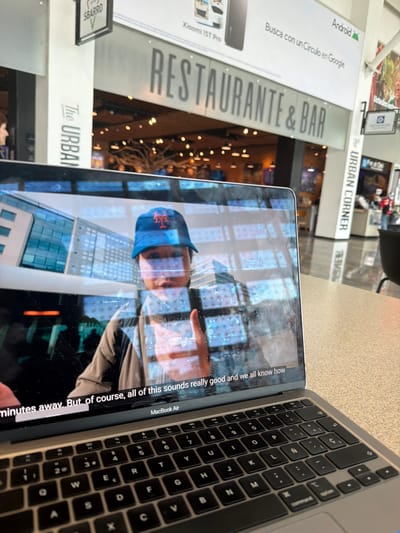 The image shows a MacBook Air laptop sitting on a table in what appears to be a public setting like a restaurant or cafe in Santa Cruz del Valle, Mexico. The screen displays a video featuring a person wearing a blue baseball cap with the New York Mets logo on it. The MacBook is placed in front of a glass wall sign for "RESTAURANTE & BAR", identified as "The URBAN CORNER." On the sign, there's a promotion for Android and Xiaomi phones. The laptop's keyboard is visible, and the screen displays subtitles. The surrounding area includes other tables, suggesting a casual dining or gathering spot.