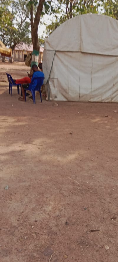 The scene is an outdoor area in Iyara, Nigeria, characterized by dry, reddish-brown earth with scattered small debris. On the right, a large, light-colored canvas tent, exhibiting a rounded top and prominent vertical seam lines, is erected. To the left of the tent, two individuals are seated on blue plastic chairs. One person, wearing a blue shirt and dark shorts, is positioned facing away from the camera, actively viewing a mobile phone held in their hands. This individual wears sandals. Adjacent, a second individual in red shorts is partially visible, reclining with their legs extended and resting on another blue plastic chair. A third person, wearing a green garment, is partially obscured behind the individual in the blue shirt. In the background, beyond several trees with sparse green foliage, are permanent structures made of stone or brick, topped with reddish-brown roofs. One tree trunk in the mid-ground is distinctly marked with green and white paint, from which a brown sack is suspended.