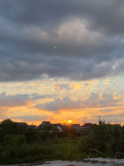 The image depicts a sunset over a rural village. The sky is filled with a mix of dark, heavy clouds and lighter, wispy clouds illuminated by the setting sun. The sun itself is partially obscured by clouds, casting a warm orange and yellow glow across the horizon. In the foreground, a cluster of houses with dark roofs are visible amongst lush green trees and vegetation. The village appears to be nestled within a natural landscape, with no signs of urban sprawl or dense population. The overall atmosphere is one of tranquility and natural beauty. The image is not located in Slobozia, Moldova, but rather appears to be a generic rural setting.