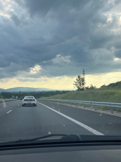 An elevated viewpoint from inside a vehicle on a multi-lane asphalt highway observes a white Ford Mustang GT traveling ahead in the right lane. The Mustang displays a Bulgarian license plate, "B 0502 HT." The road features clear white dashed lane markings and a solid white line on the right shoulder. A metal guardrail parallels the right side of the highway, beyond which is a grassy embankment with green vegetation and sparse trees. A prominent metal lattice tower stands beside a leafy tree on the right side, further from the road. In the distance, a range of hills or mountains stretches across the horizon, with indistinct urban structures visible in the intervening plain. The sky is predominantly overcast with dark, heavy clouds, but rays of sunlight penetrate the clouds, illuminating parts of the landscape, particularly towards the horizon on the right. The view from the perspective vehicle's dashboard, including windshield wipers, is visible at the bottom of the frame. The scene is consistent with a highway approach or departure from Sofia, Bulgaria, near Dolni Bogrov.