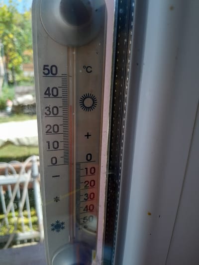A close-up, eye-level, vertical shot shows a thermometer mounted on a window frame. The thermometer has a scale marked in degrees Celsius, with visible numbers ranging from 0 to 50. A red liquid column indicates the temperature, which appears to be around 26 degrees Celsius. The background, visible through the window, shows a blurred outdoor scene with greenery and trees under a bright sky. The window frame is white, and part of another window or wall is visible on the right side. The thermometer itself is white and slightly dusty.