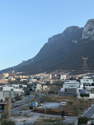 A stark mountain range dominates the left of the frame under a clear sky. Nestled along the base of the mountain are tightly packed houses, appearing in shades of white and beige. A power line tower is visible on the mountain slope. A road winds through the residential area, bordered by vegetation.