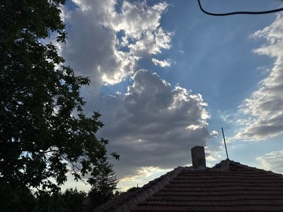 The image depicts a view of the sky over rooftops in Levski, Bulgaria, likely during the late afternoon or early evening given the lighting. A large tree with dark green foliage dominates the left side of the frame, its branches reaching upwards towards the sky. In the background, a tiled roof of a building is visible, characterized by its reddish-brown tiles and rough cement finishing. A chimney made of concrete blocks stands on the roof, and a thin black antenna or rod extends vertically from another point on the roof. The sky is a dramatic mix of blue and grey clouds. Some clouds are illuminated by sunlight, appearing bright white and fluffy, while others are darker and more textured, suggesting impending weather changes or the setting sun. The overall scene is one of a residential area under a dynamic sky.