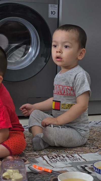 This is a portrait-oriented, vertical media file showing two young children in an indoor setting in Mary City, Turkmenistan. The primary subject is a toddler boy in the foreground, wearing a grey t-shirt with red lettering that appears to say "TOMMY HILFIGER" and a German flag emblem. He is looking towards the camera with an open mouth and wide eyes. He is sitting on a patterned rug next to a large, dark grey washing machine. A second child, dressed in a red outfit with orange polka dots, is partially visible in the lower right corner, also sitting on the rug.
The setting appears to be a home, with a patterned rug covering the floor and what looks like a table runner or tablecloth with text on it in the background. The visible text on the tablecloth includes "COFFEE" and "EVERY DAY". There are also two bowls, one blue and white and the other white, containing a light yellow liquid, presumably some kind of drink. An orange-handled utensil, possibly a spoon or a spatula, is also present on the tablecloth.
The overall mood is casual and domestic, with the children engaged in their own activities. The lighting suggests it could be daytime. No specific emotions are strongly conveyed beyond the child's curious expression. The presence of a washing machine and everyday household items like bowls and utensils suggest a lived-in environment.