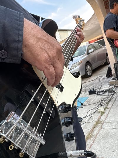 The image displays an outdoor musical performance taking place in Chamilpa, Mexico. In the foreground, a person's hands are actively playing a black electric bass guitar, which features a white pickguard, four strings, a chrome bridge, two black pickups, and three black control knobs. A silver audio cable is plugged into the instrument's input jack. The left hand with visible wrinkles frets notes on the light-colored fretboard, while the right hand plucks the strings. The musician wears a dark, long-sleeved shirt or jacket, with a black button visible on the cuff. To the right and slightly in the background, another individual is partially visible, wearing a dark t-shirt and holding a red electric guitar. The setting is outdoors on a light-colored concrete or paved surface, where a silver four-door sedan is parked behind the musicians. Above the vehicle, a light-colored arched architectural element of a building is visible, alongside a partial awning or roof structure. Numerous black and white electrical cables, power strips, and small black electronic devices are spread across the ground. The sky is clear blue with minimal cloud cover.