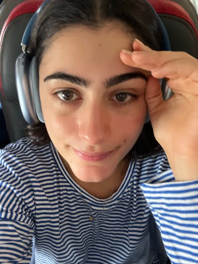 A close-up, high-angle, selfie-style shot features a young woman in Paray-Vieille-Poste, France. She is wearing blue and white striped long-sleeved shirt and large, light blue over-ear headphones. Her right hand is raised and touching her forehead and temple, obscuring the right side of her face. Her left eye is visible and looking directly at the camera. The background consists of the dark grey and red upholstery of a chair.