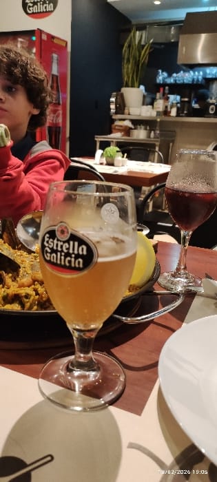 A table setting features a glass of "Estrella Galicia" beer and a glass of red wine. A plate sits above them. The beer glass is partially filled. Below, a pan contains food, possibly seafood-based. A child in a red shirt appears in the corner. The backdrop includes a refrigerator with "Estrella Galicia" branding and a glimpse of a bar-like setting with plants.