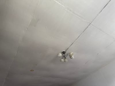 The image shows a close-up, low-angle view of a white, textured ceiling in a room. There are visible seams and imperfections on the ceiling, suggesting it may be made of plasterboard or a similar material. A single, multi-bulb chandelier hangs from the center of the ceiling, its metallic fixture and glass shades reflecting the light. The chandelier appears to be old-fashioned, with a somewhat ornate design. The room appears to be indoors, and the lighting suggests it is daytime, though no windows or external views are visible. The overall impression is of a simple, possibly older room in Russia, given the location context of Pozdyshki. There is a small, circular stain on the ceiling to the left of the chandelier, possibly indicating a water leak. The texture of the ceiling is uneven, with small bumps and irregularities. No people or text are visible in the image.