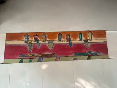 The image shows a colorful, abstract painting mounted on a white tiled wall. The painting, which appears to be abstract, features several stylized vase-like shapes in various colors, including gray, brown, red, and green, arranged in a row across an orange and tan background. Below this row, a vibrant pinkish-red area represents water, with several boat-like shapes depicted in blues, greens, yellows, and browns. The painting is long and narrow, suggesting it might be a mural or a long decorative piece. The white tiled wall suggests an indoor setting, possibly a kitchen or bathroom given the tiled surface. The lighting in the image creates shadows on the lower part of the wall, indicating ambient light from an unseen source. The specific location of Karangkebagusan, Indonesia, is mentioned, but the image itself provides no visual cues to confirm this geographical detail.