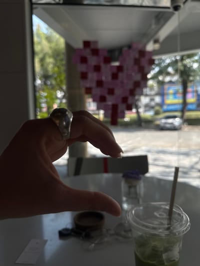 A hand with a large silver ring is in the foreground. In the background, visible through a window, is an abstract pixelated art piece in pink and red hues and greenery. A clear plastic cup containing a green beverage with a straw sits on a white surface, along with a small, dark-colored container and what appears to be a folded card. A reflective surface adds to the complexity of the composition.