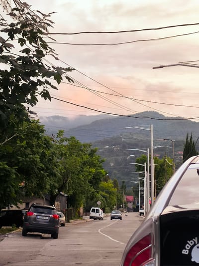 The image depicts a street scene in Khelvachauri, Georgia, with a focus on the natural landscape and the urban environment. In the foreground, the back of a silver car is visible, with a "Baby on Board" sticker on its rear window. The car is parked on a street that shows signs of wear, with visible cracks and uneven surfaces. Adjacent to the street, dense green foliage from trees lines the left side of the frame, with branches extending towards the top. Beyond the trees and the street, a series of modern streetlights stand tall, casting shadows. In the midground, several vehicles are present, including a dark gray SUV with license plate BA-262-BU, a white van, and a silver sedan. The road curves slightly to the right, leading into the distance. The background reveals rolling green hills and mountains, partially obscured by a soft, misty haze. The sky above is a pale pink and orange hue, suggesting either dawn or dusk, with a network of power lines crisscrossing the frame. Scattered houses and buildings are visible nestled on the slopes of the hills, indicating a residential area integrated with the natural environment. The overall atmosphere is serene and captures the transition between a developed area and the natural beauty of the Georgian countryside.
