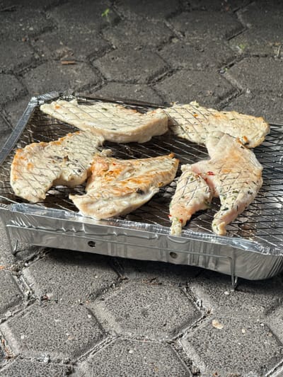 This image depicts five pieces of marinated chicken breast cooking on a portable grill. The grill is a disposable aluminum foil tray with a wire rack. The chicken pieces are arranged on the rack, some showing light browning and grill marks. The grill is placed on a paved surface, possibly a patio or sidewalk, made of gray interlocking bricks. The background shows more of the paved area with some dirt and debris. The overall scene suggests an outdoor cooking activity. The image was taken in Sankt Wendel, Germany.