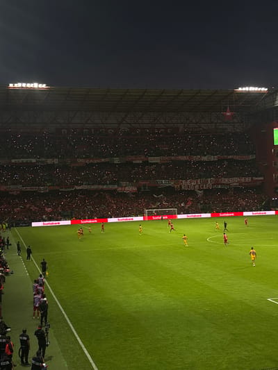 The image shows a night-time soccer game in Toluca, Mexico. The stadium is packed with fans, many holding up lights, creating a luminous display in the stands. Banners in Spanish are visible among the crowd. On the field, players in red and yellow uniforms are engaged in the game. Advertisements for Scotiabank line the perimeter of the field. The stadium lights illuminate the scene. A white line demarcates the edge of the field. Camera operators are positioned along the sidelines. The scene conveys a vibrant atmosphere, typical of a major soccer match.