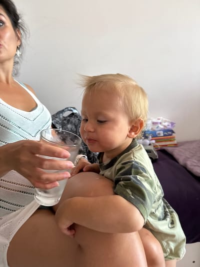 In this indoor, natural light photograph taken in Sochi, Russia, a young toddler with blond hair leans forward, looking down with interest. The toddler is wearing a camouflage t-shirt and is positioned on top of a woman's torso. The woman, with dark hair and wearing a light blue, lacy top, is partially visible from the chest up. She holds a clear glass in her right hand, which appears to contain a liquid. Her left hand is also visible. The background is blurred, showing hints of a wall and some colorful objects, possibly books or toys, stacked on a surface. The overall scene suggests a moment of interaction between the child and the woman, perhaps during feeding or play. There is no visible text in the image. The emotions conveyed are one of curiosity and gentle engagement.