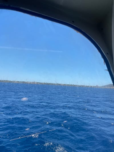The image presents an outward view from inside a vessel or low-flying aircraft, overlooking a vast expanse of deep blue water with a distant coastline visible under a clear, bright blue sky. The top and right portions of the frame reveal the light-colored, curved interior structure of a window or cockpit, secured by visible fasteners along the right edge.
The sky occupies the upper third of the image, devoid of clouds, with a single faint, horizontal trace line apparent. The majority of the frame is dominated by the water, which exhibits moderate surface ripples and small waves. Submerged or semi-submerged objects are discernible in the water; these include several indistinct, elongated light-colored forms and a horizontal, linear chain-like object in the lower-left quadrant.
The distant coastline, identified as part of Marsala, Italy, spans the horizon. It features a continuous strip of structures, including buildings with varied rooflines and some interspersed greenery, suggesting a developed coastal area. The land gently rises towards a small headland or hill on the far right. No human figures or discernible specific actions are present within the frame. The scene is illuminated by direct daylight.