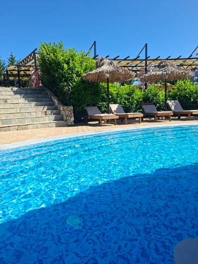 The image depicts an outdoor swimming pool with clear blue water, tiled in a mosaic pattern of various shades of blue. Sunlight reflects off the water's surface, creating ripples and highlights. Along the edge of the pool, a paved walkway leads to a set of stone stairs. Behind the pool and stairs, a lush green hedge forms a backdrop. Several thatched-roof umbrellas and lounge chairs are arranged in a seating area beneath a pergola structure. The sky above is a clear, bright blue. The scene is set at the Ravadinovo castle complex in Ravadinovo, Bulgaria. There are no people visible in the image.