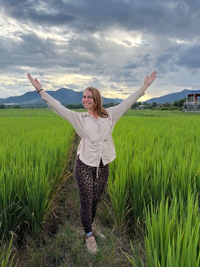 A woman stands with her arms outstretched in a vibrant green rice paddy field. She is wearing a light-colored button-down shirt with cinched waist and leopard-print leggings. She has light brown hair and is smiling at the camera. The rice field stretches out behind her, with mountains visible in the distance. The sky is partly cloudy, with hints of sunlight breaking through. The scene appears to be located in the city of Ban Pong, Thailand, as indicated by the landscape.