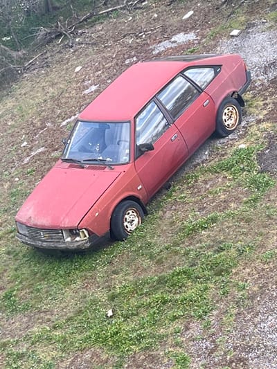 A red, older-model hatchback, consistent with a Moskvitch 2141 "Aleko", is observed parked or stranded on a sloped, uneven terrain in Bolyarovo, Bulgaria. The vehicle is oriented facing downhill, with its front end directed towards the lower part of the slope. The car's paint is faded and appears dirty. The front bumper exhibits damage, with the headlight assembly on the viewer's left (passenger side) missing or severely compromised. The front hood shows a slight misalignment. All four wheels are present, featuring off-white, possibly steel, rims stained with rust and dirt. The surrounding environment consists of patchy green grass intermingled with areas of exposed soil and gravel, indicating a rugged, unpaved surface. Further up the slope in the background, denser vegetation, including trees or shrubs, and scattered dry branches are visible. No individuals or animals are present in the scene, and no actions or interactions are depicted.