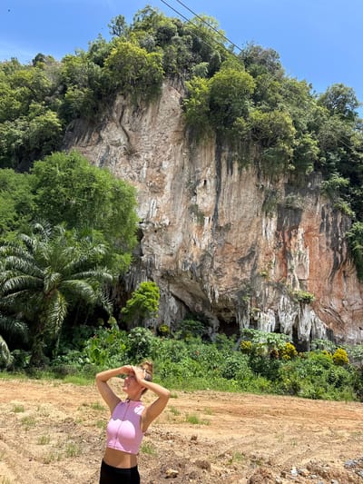 A young woman with blonde hair in a bun, wearing a pink crop top and black bottoms, stands in a dirt clearing. She is looking up and to her left, shielding her eyes with her hands, appearing to be squinting at the bright sun. Behind her, a towering limestone cliff face is covered in lush green vegetation, with visible stalactites hanging from its overhangs. The cliff also shows streaks of reddish-brown and gray mineral deposits. In the foreground, the ground is a mix of dry dirt and sparse green grass. The sky above is a clear, bright blue. The scene is set in a natural, tropical environment, likely in the Thung Yai region of Thailand, known for its karst topography.