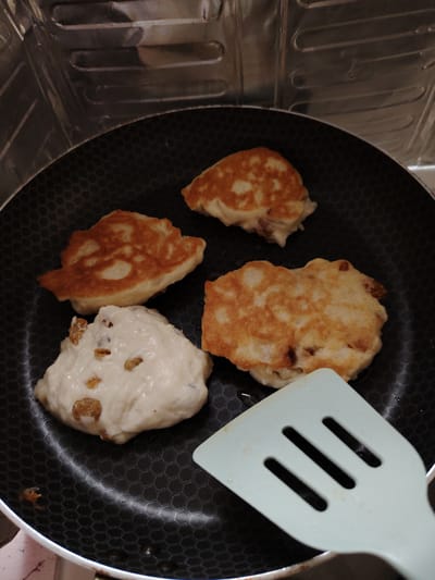 In Soivolovskaya, Russia, an overhead shot captures four pancakes cooking in a black hexagonal-patterned non-stick pan. Three of the pancakes are golden brown, indicating they are nearly done or have been flipped, while one pancake remains pale and appears to be raw batter with visible raisins. A pale blue spatula with three slits is positioned at the bottom right of the pan, partially obscuring one of the cooked pancakes. The background is a blurred metallic surface, possibly a stove or backsplash, reflecting light. The lighting suggests indoor cooking conditions, likely in a domestic kitchen.
