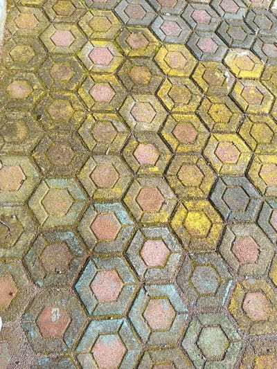 The image displays an overhead view of an outdoor paved surface located in Salai Har Htan, Myanmar. The pavement consists of numerous interlocking hexagonal concrete pavers arranged in a tessellated pattern. Each paver unit features a raised perimeter and a central hexagonal depression. The pavers exhibit a weathered appearance with a varied color scheme, including faded red-pink, light grey, and yellow hues. Extensive organic growth, consistent with moss or algae, is present as mottled green and yellowish discoloration covering large areas of the pavers and their interstices. Faint patches of light blue pigment are visible on some paver sections. The joints between the pavers are filled with loose dirt, small gravel, and organic debris. No individuals, objects other than the paving material, or actions are depicted within the scene.